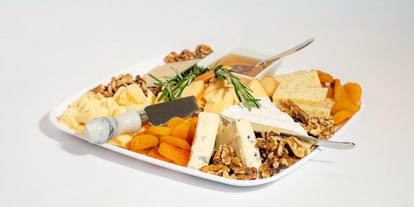 A cheese platter with dried apricots, walnuts, honey, and herbs on a white tray.