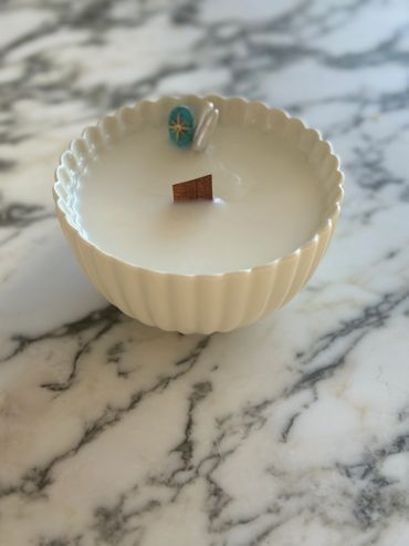 White scalloped bowl candle with wooden wick on marble surface.