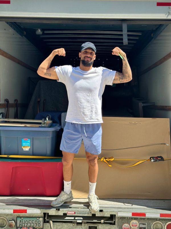 Man in white t-shirt and blue shorts flexing muscles in a moving truck.