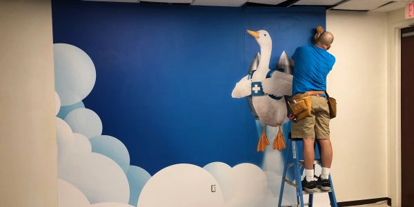 Man installs a mural of a medical goose flying among clouds on a blue wall.