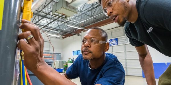 Two men wearing safety glasses work on a mechanical project indoors.