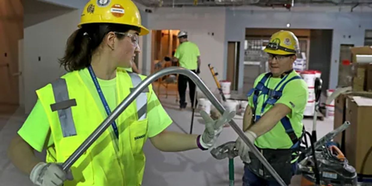 Two construction workers in safety gear handling a bent metal pipe indoors.