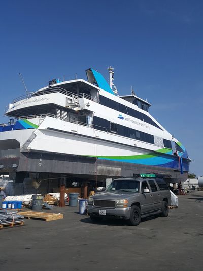 Large San Francisco Bay Ferry vessel out of the water on supports, with a silver SUV parked nearby.