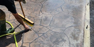 Person cleaning patterned concrete with broom and hose.