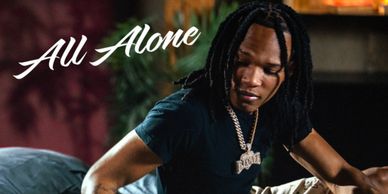 Man with dreadlocks wearing a chain and dark shirt with 'All Alone' text.