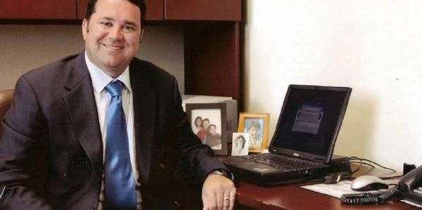 A man in a suit smiling at his office desk with a laptop and framed photos.