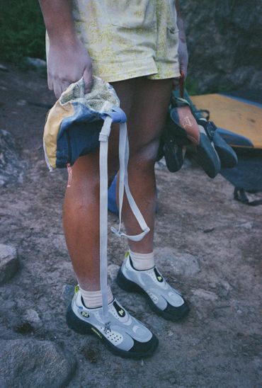 A person holding climbing shoes and chalk bag, wearing climbing shoes.