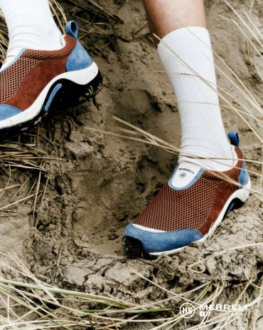 Person wearing Merrell shoes standing on sandy ground with grass.