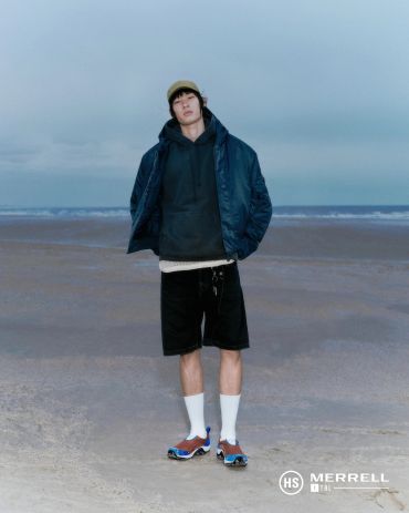 Young man in casual sporty outfit standing on a beach under cloudy sky.