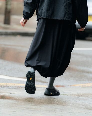 Person walking on wet pavement wearing black shoes and a long black skirt.