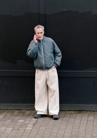 Man in a green jacket and wide beige pants talking on a phone.