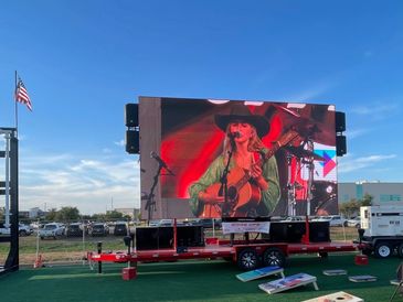 Outdoor event with a large screen showing a woman playing guitar and cornhole boards on green turf.