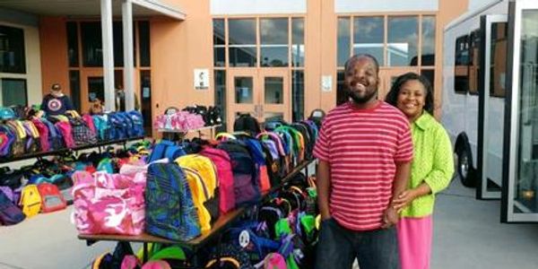 Couple stands smiling by tables full of colorful backpacks outside a building.