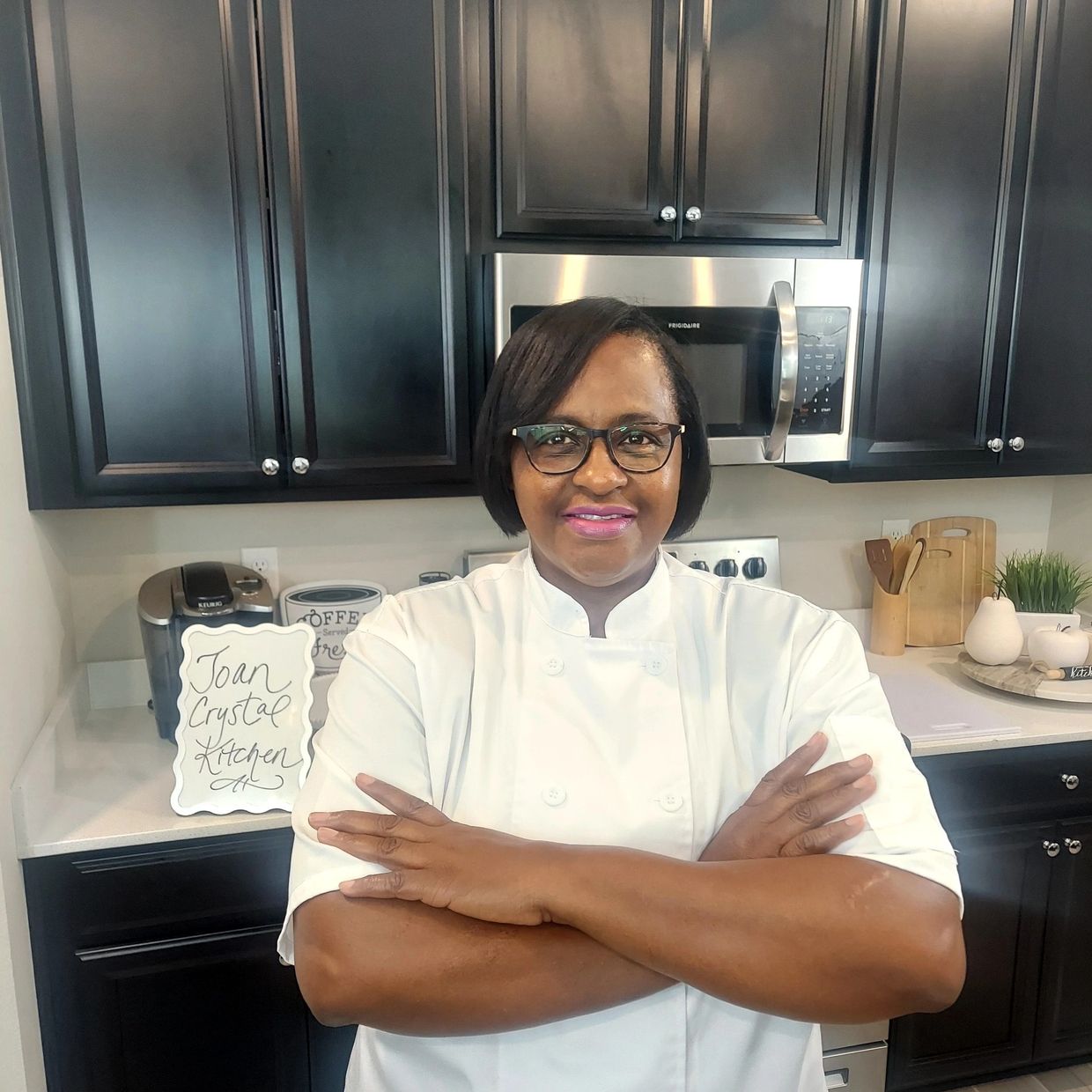Confident chef in white uniform standing in a modern kitchen with crossed arms.
