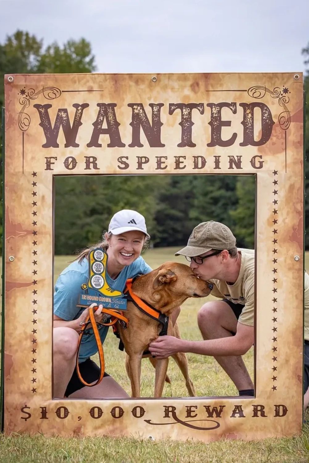 Two people posing with a dog in a "Wanted for Speeding" frame outdoors.