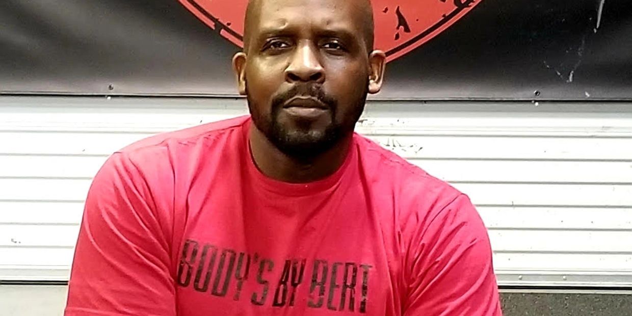 Man wearing a red 'BODY'S BY BERT' t-shirt posing indoors.