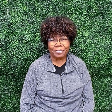 Smiling woman with curly hair and glasses in front of a green leafy wall.