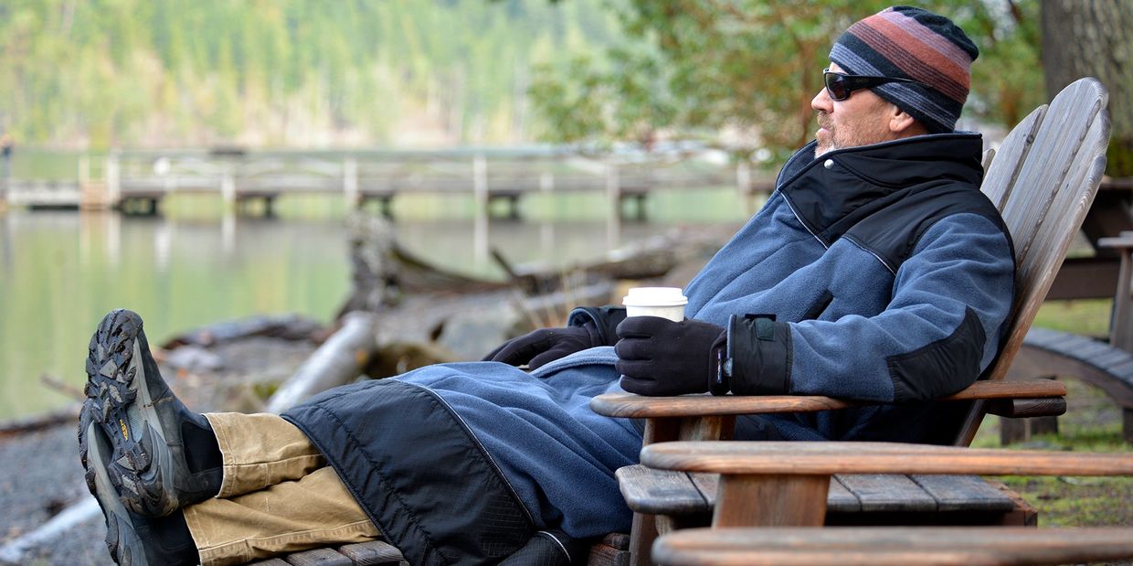 man lounging in a rugged robe at a lake drinking coffee