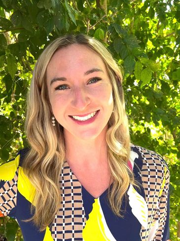 Smiling woman in patterned blouse outdoors with greenery.