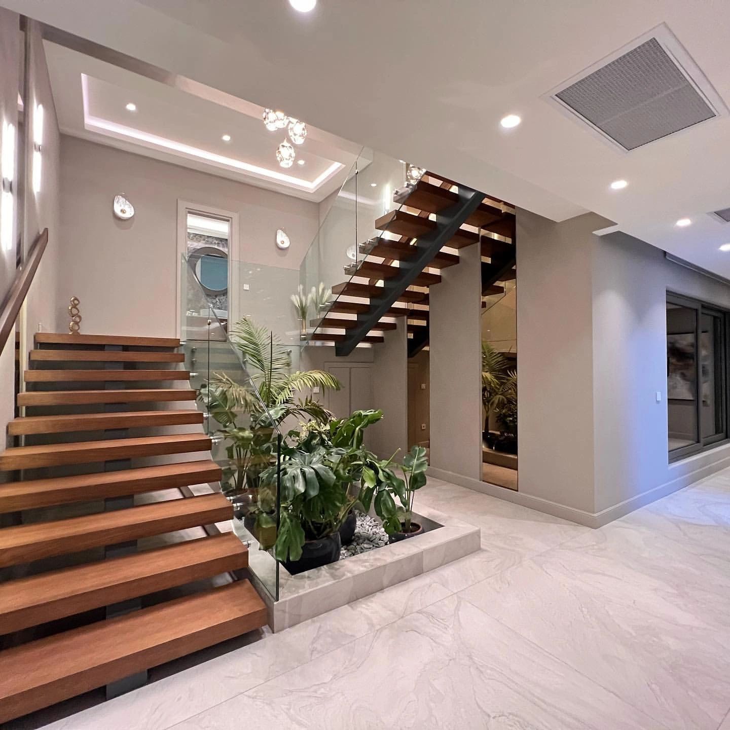 Modern wooden staircase with glass railing and indoor plants in a sleek foyer.