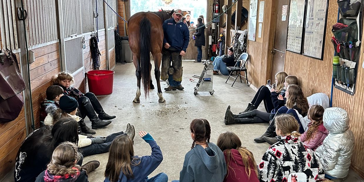 Children sitting in a stable observing a farrier work on a horse's hoof.