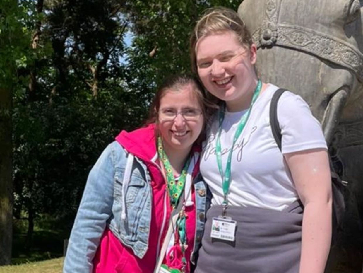 Two women smiling outdoors on a sunny day, standing close to each other.