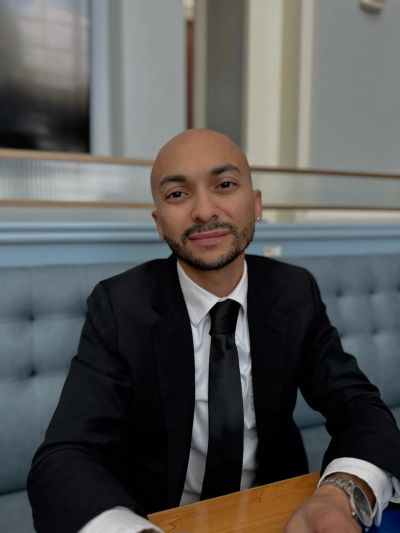 Man in a black suit and tie sitting at a table, smiling.