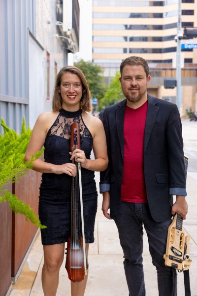 Two musicians posing with string instruments on a city sidewalk.