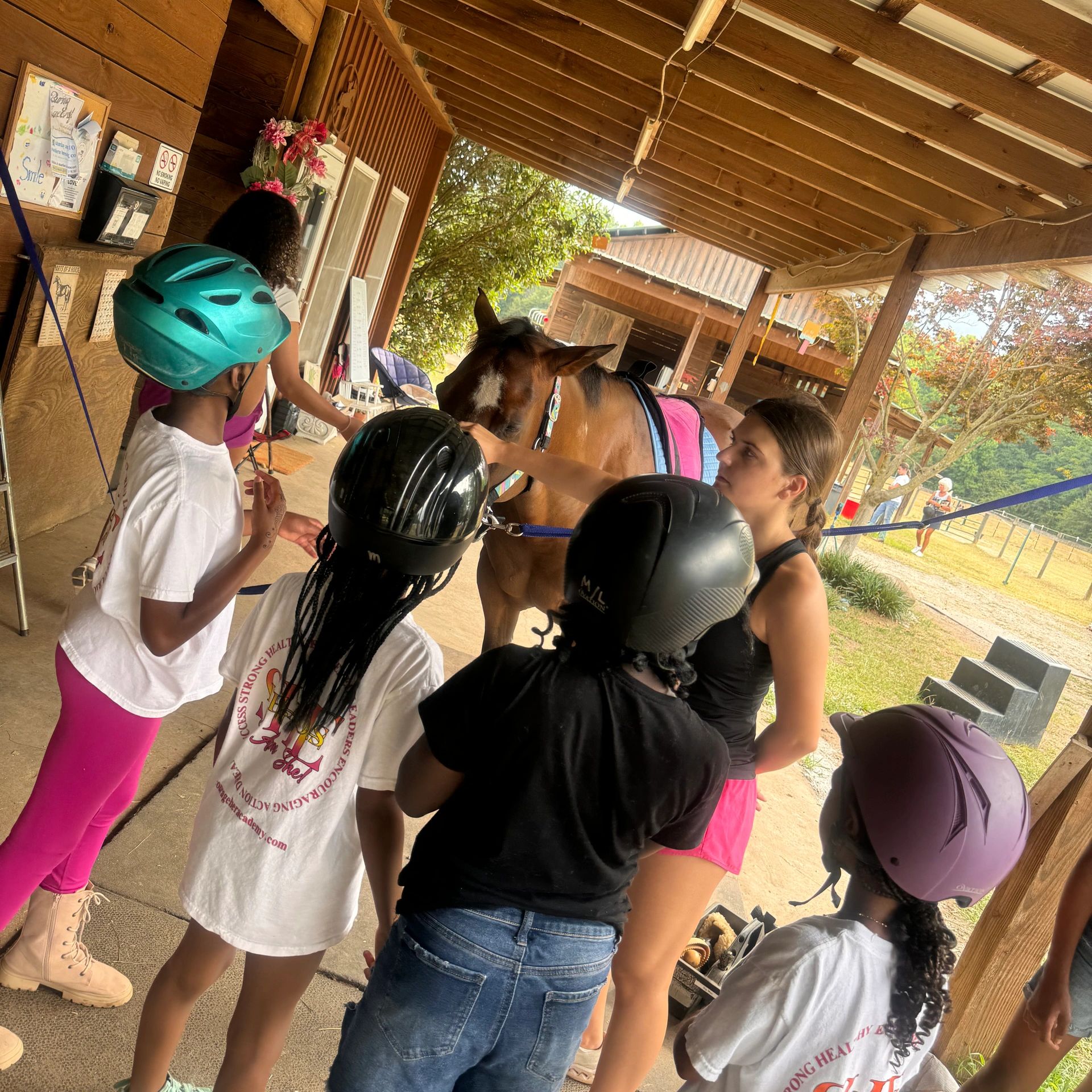 Kids wearing helmets gather around a horse under a wooden shelter.
