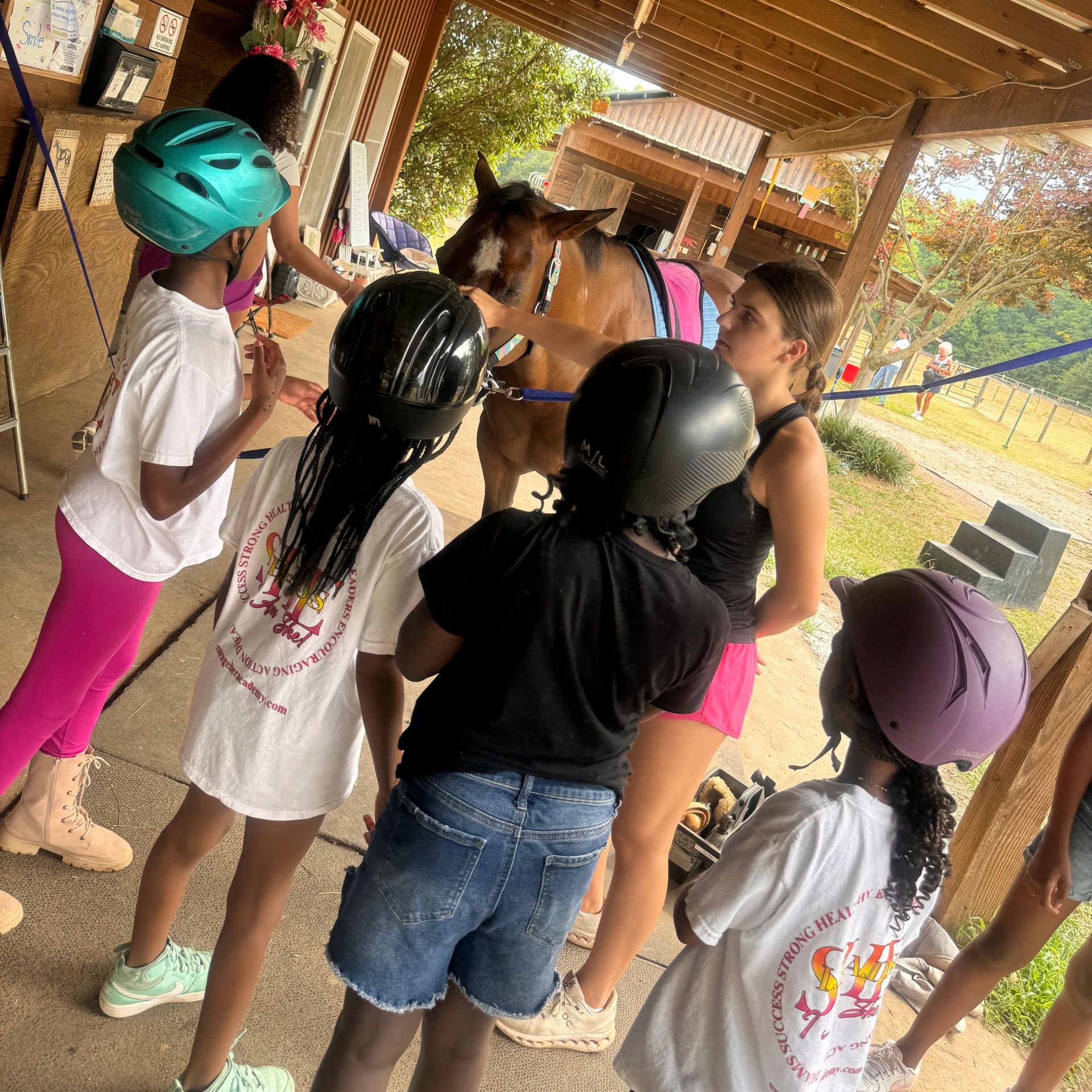 Kids wearing helmets gather around a horse under a wooden shelter.