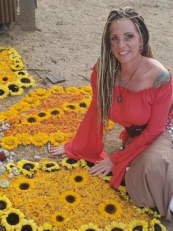 Woman in red top with dreadlocks kneeling by sunflower petal art outdoors.