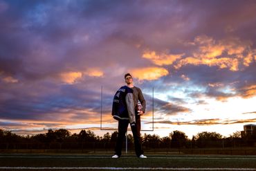 Young man with football and letterman jacket at sunset on field.