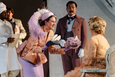 A woman in vintage attire offers a bouquet to a seated lady during a theatrical scene.