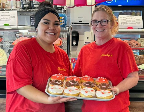 Two women in red shirts proudly hold a tray of decorated donuts at a bakery.