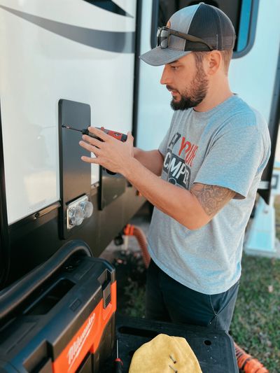 Man using a power drill on an RV exterior panel outdoors.