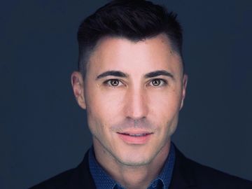 Close-up portrait of a confident man in a dark blazer and blue shirt against a dark background.