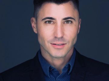Close-up portrait of a confident man in a dark blazer and blue shirt against a dark background.