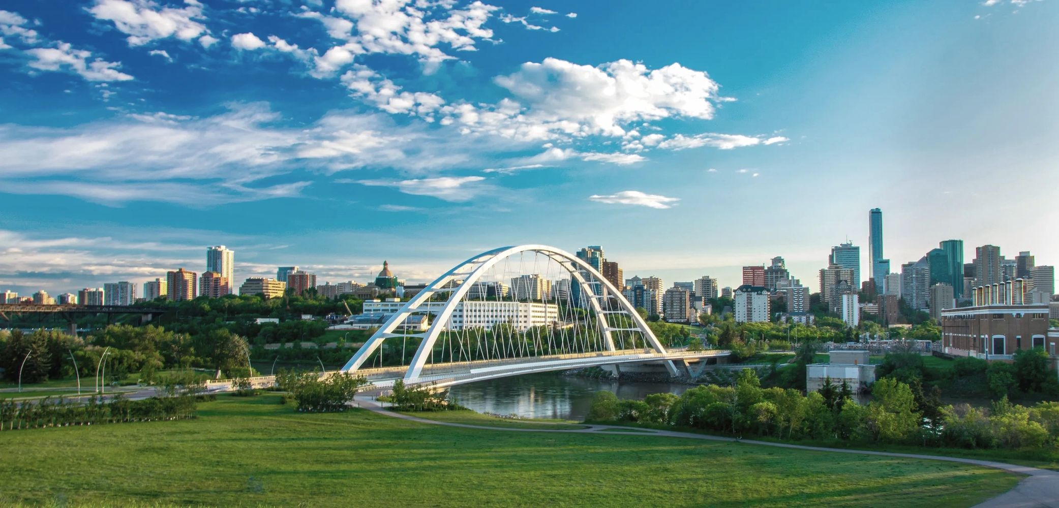 City skyline with a white arched bridge over a river under a partly cloudy sky.