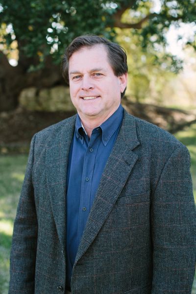Man in a blazer and blue shirt smiling outdoors with trees in background.
