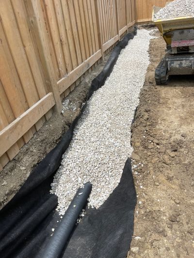 Gravel-filled trench with drainage pipe along wooden fence under construction.