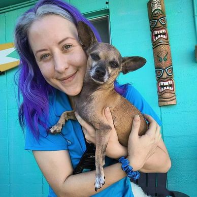 Young woman with purple hair holding a small dog outdoors.