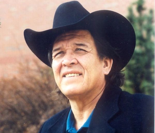 Man wearing a black cowboy hat and dark jacket looks upward outdoors.