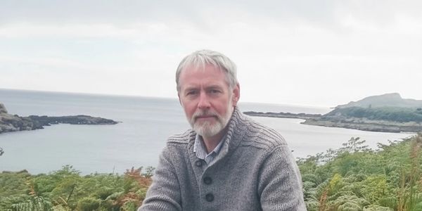 A man with gray hair and beard sits outdoors near a coastal area.