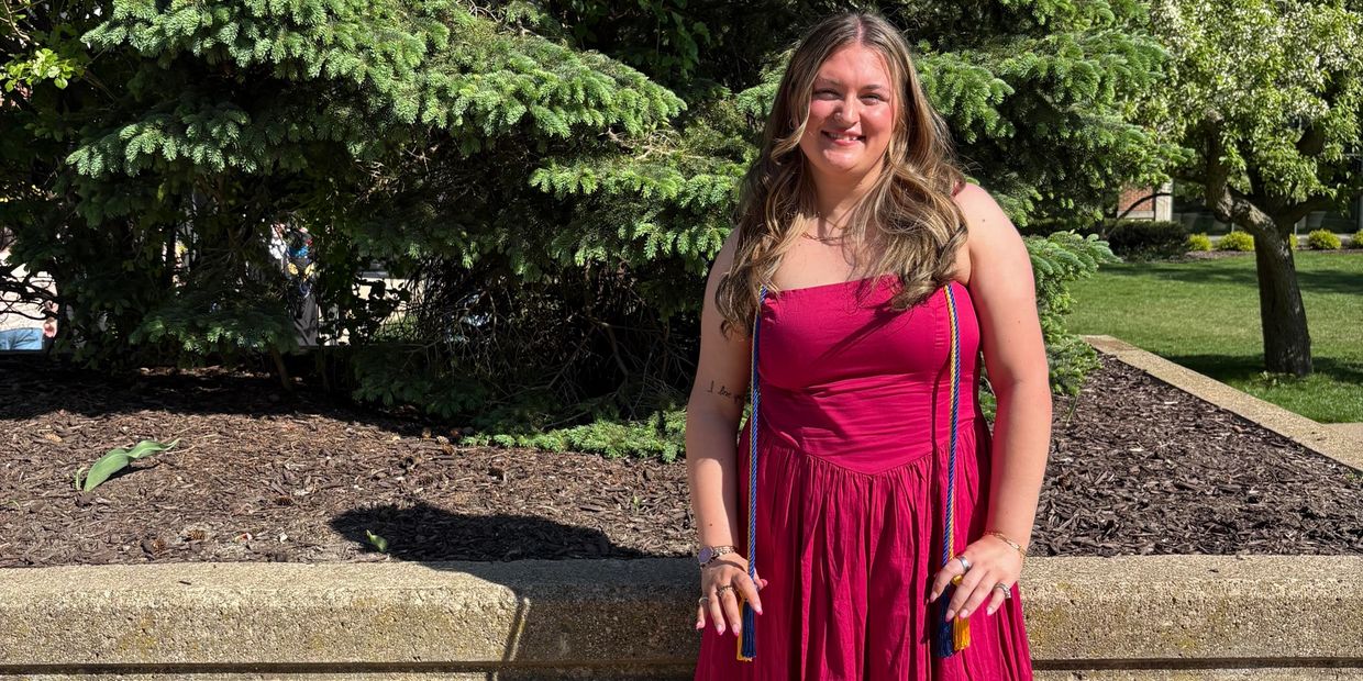 A smiling woman in a red dress stands outdoors near greenery and a concrete ledge.