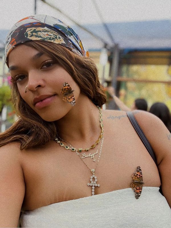 Young woman with butterflies on her face and shoulder, wearing layered necklaces and a colorful headscarf.