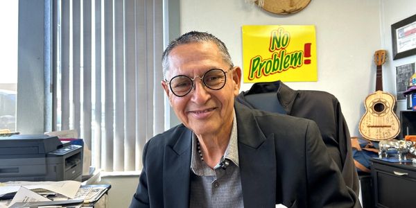 An elder person sitting in an office wearing black suit.