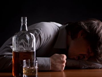Man with head down beside a bottle and glass of whiskey on a table.