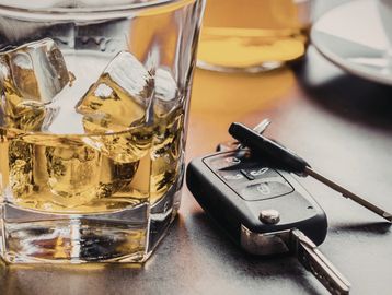 Glass of whiskey with ice and car keys on a table.