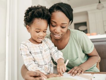 A mother helps her child with drawing or writing at a table.