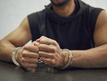 Man in black sleeveless hoodie with hands cuffed on table.
